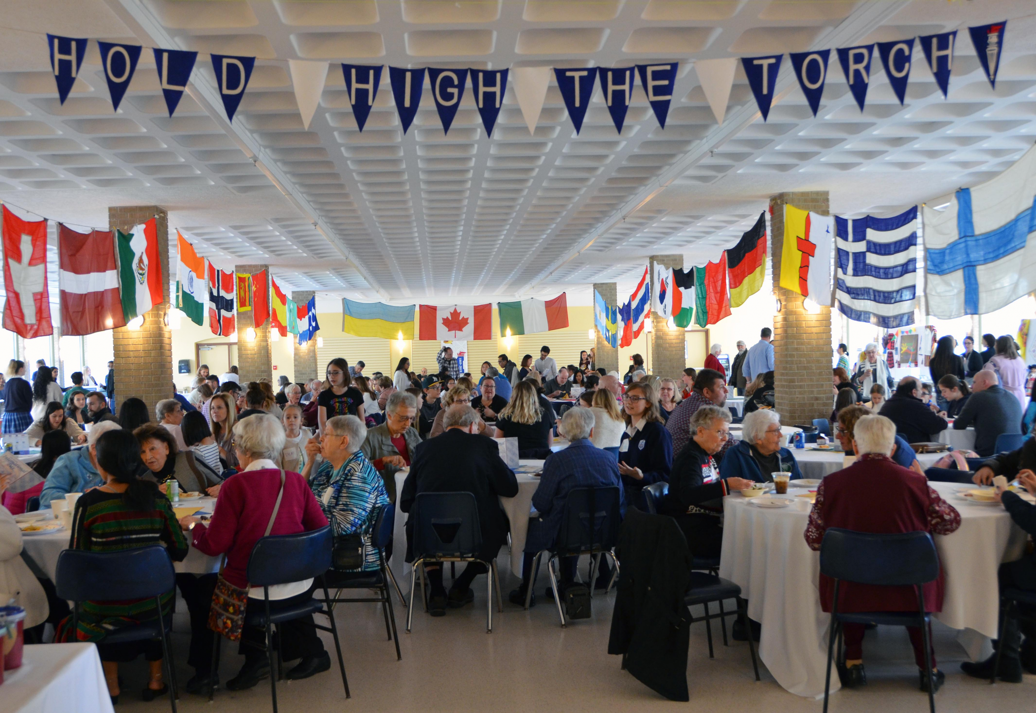 A full room of guests sit at tables during St. Mary's Academy International Day, with flags from around the world hanging from the ceiling.