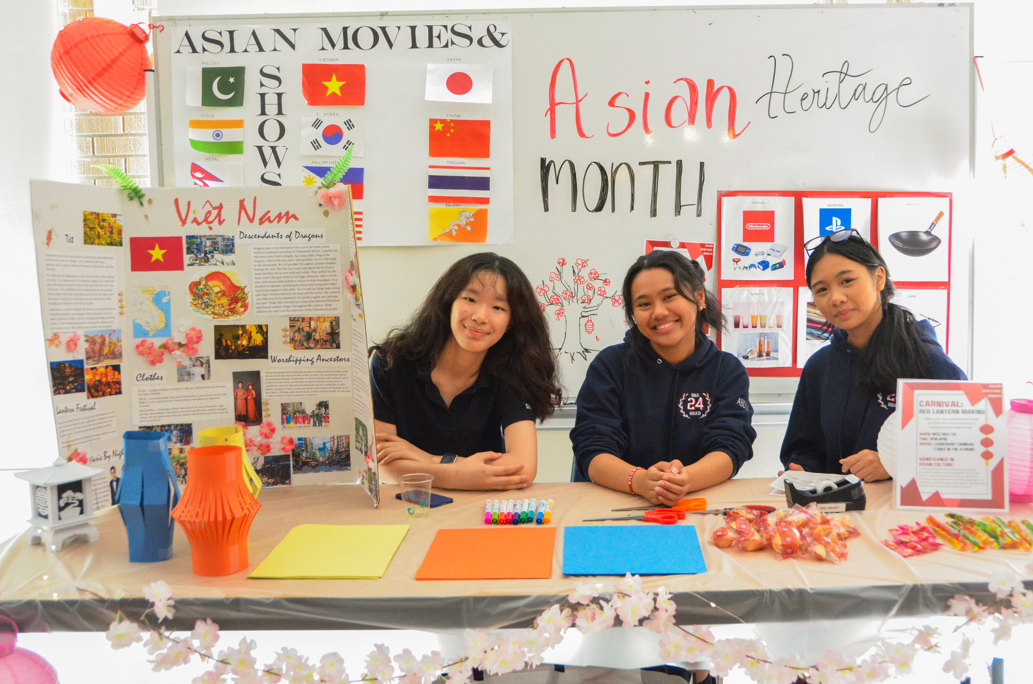 Three smiling St. Mary's Academy students sit at a table with 'Asian Heritage Month' on a poster behind them.