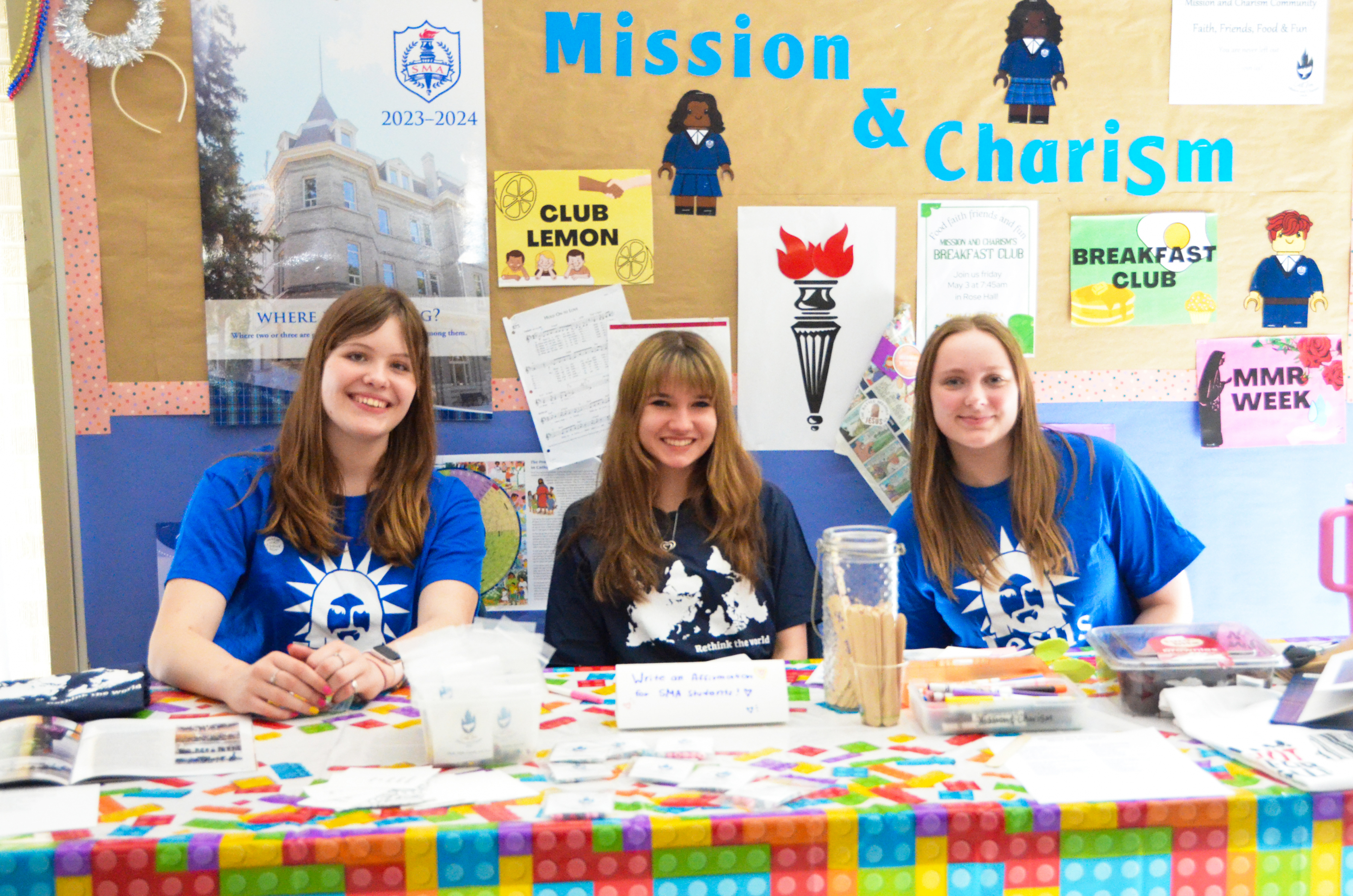 Three smiling St. Mary's Academy students sit at a table with 'Mission & Charism' and various items on a board behind them.