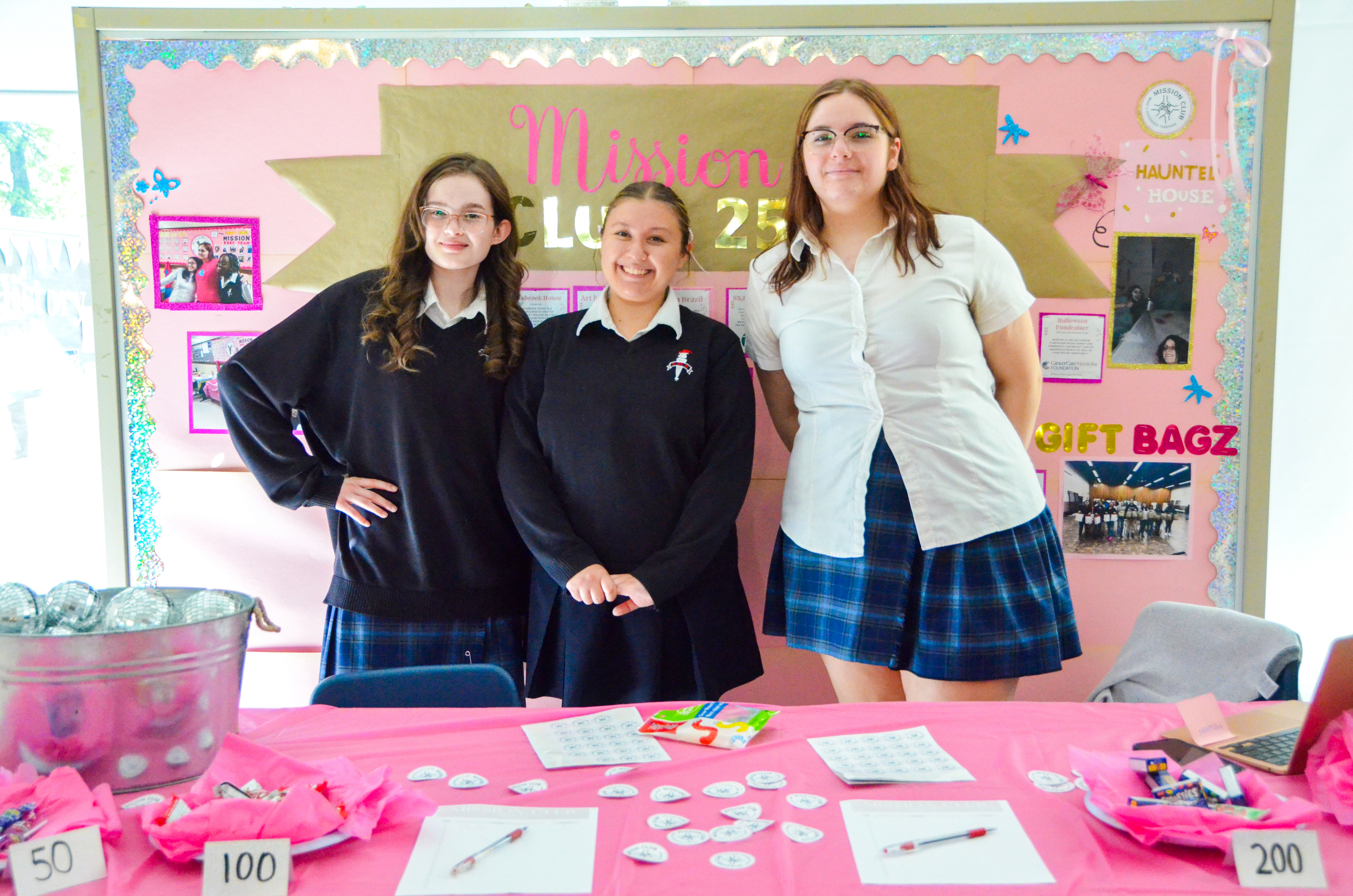 Three smiling St. Mary's Academy students stand behind a table with a pink tablecloth on it.