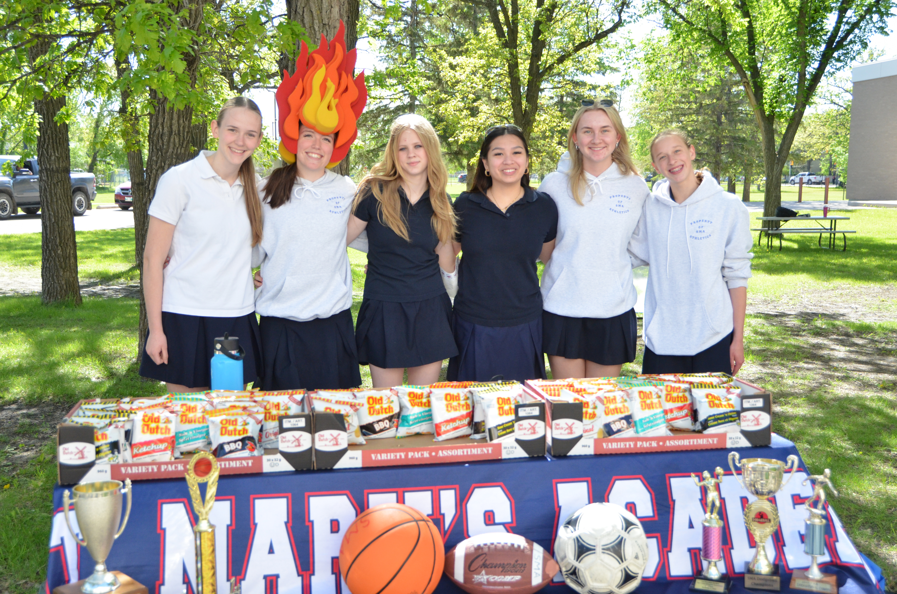 Six St. Mary's Academy students stand behind a table with various sports balls in front of it and Old Dutch chips on it.