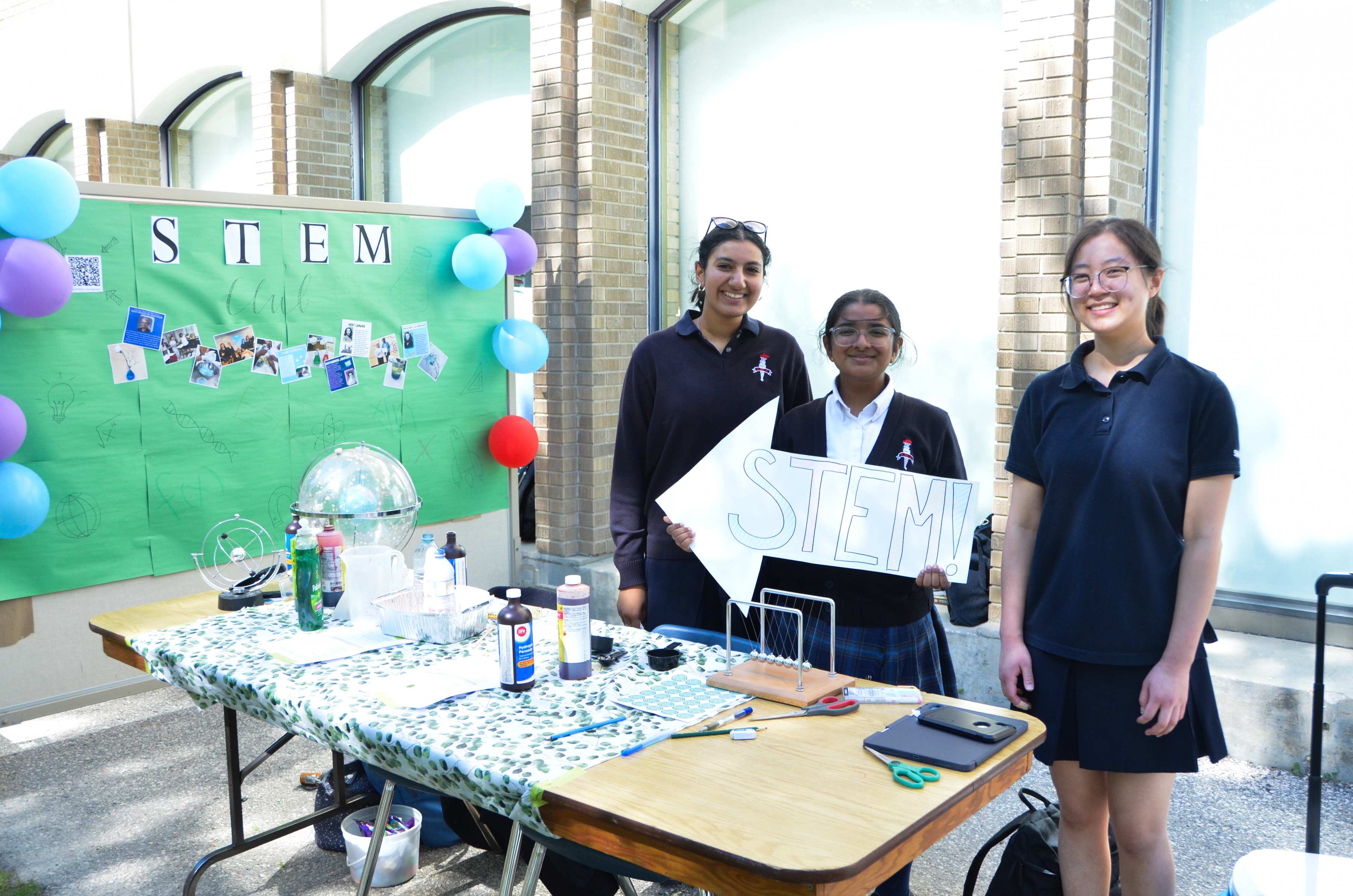 Three smiling St. Mary's Academy students stand behind a table, with one holding a sign that says 'STEM' and a poster with 'STEM Club' on it behind them.