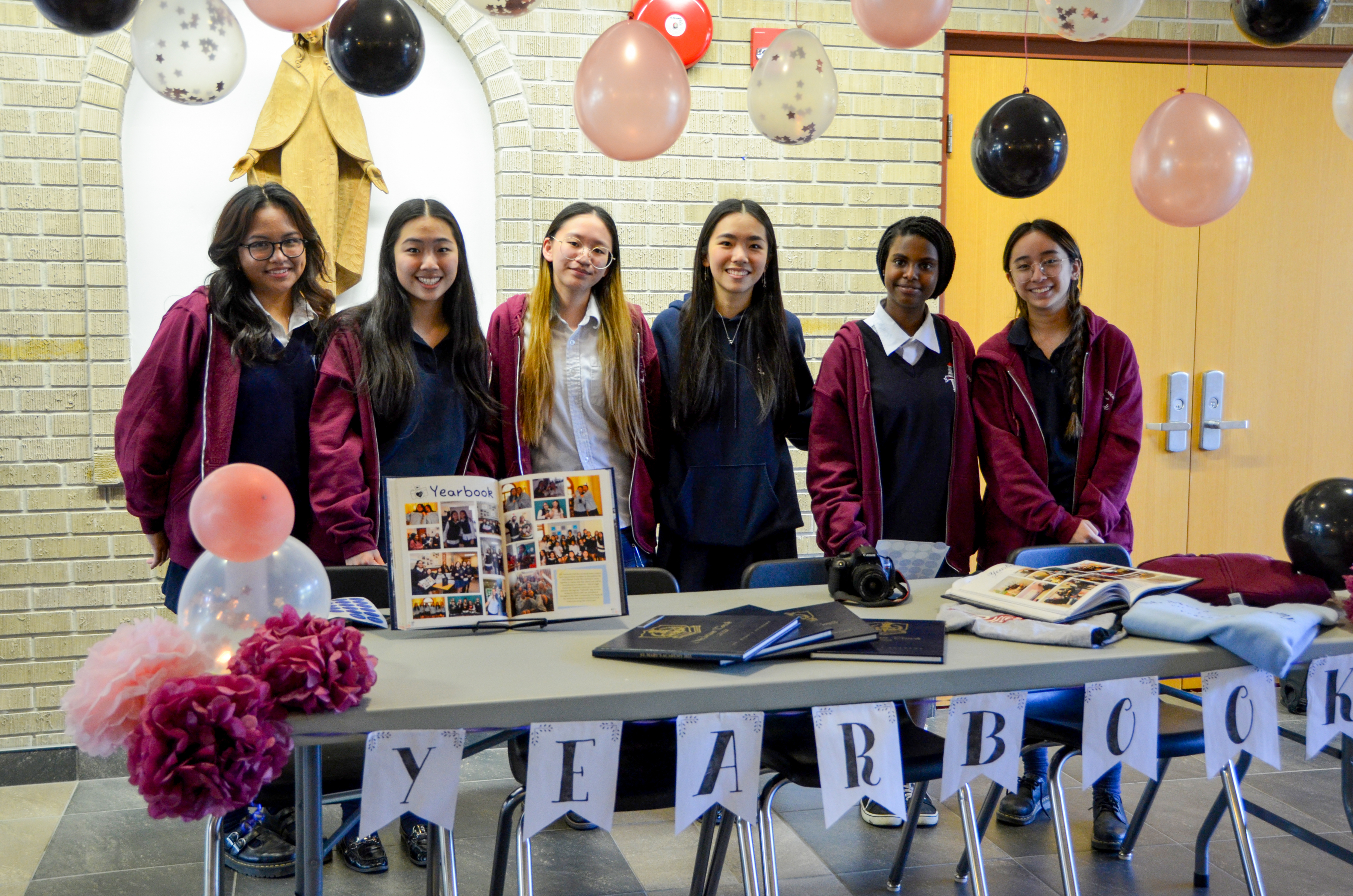 Six smiling St. Mary's Academy students stand behind a table with banners reading 'Yearbook' hanging off it and balloons above them.