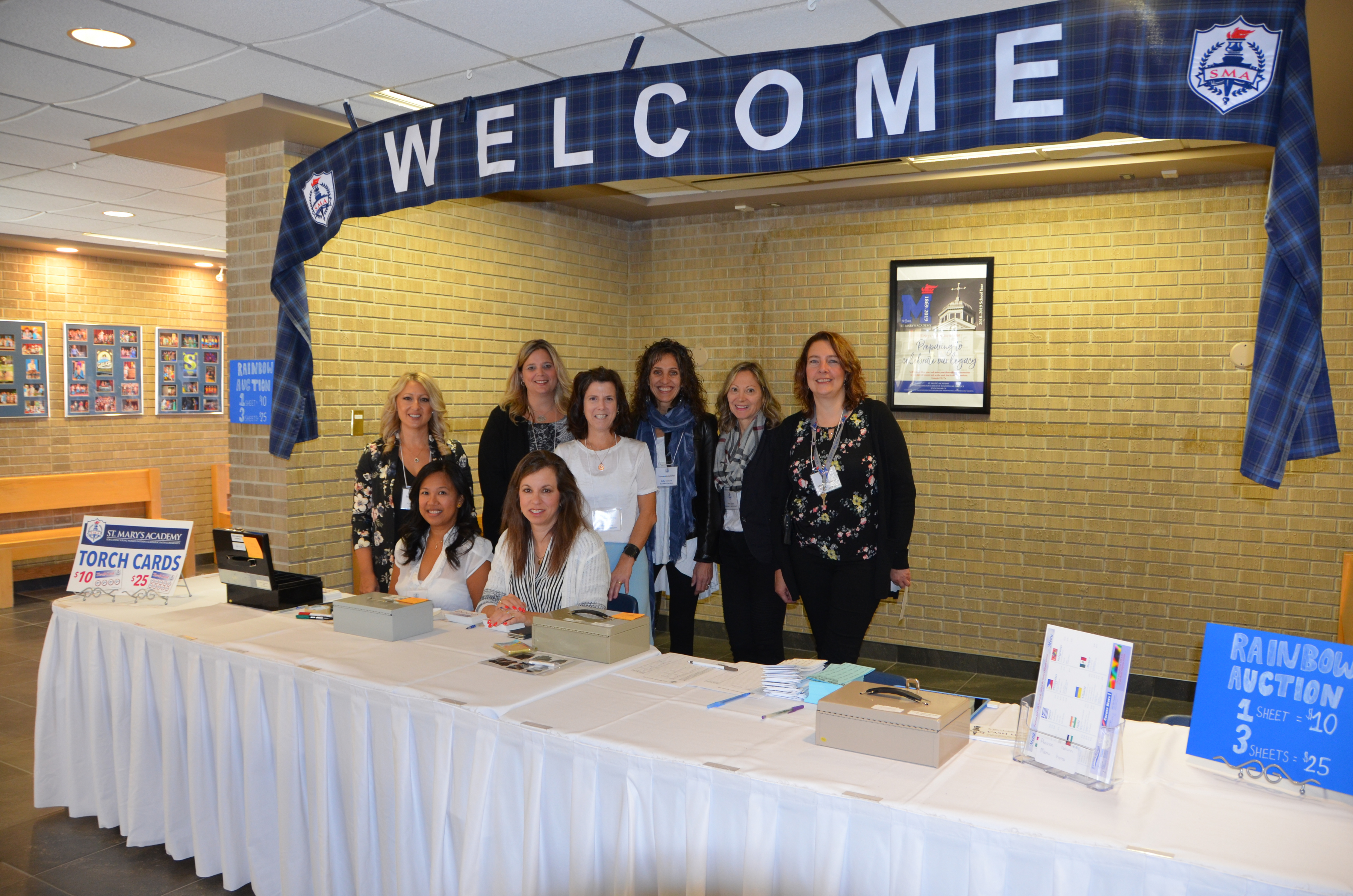 A group of eight adults stand under a Welcome banner at St. Mary's Academy International Day.