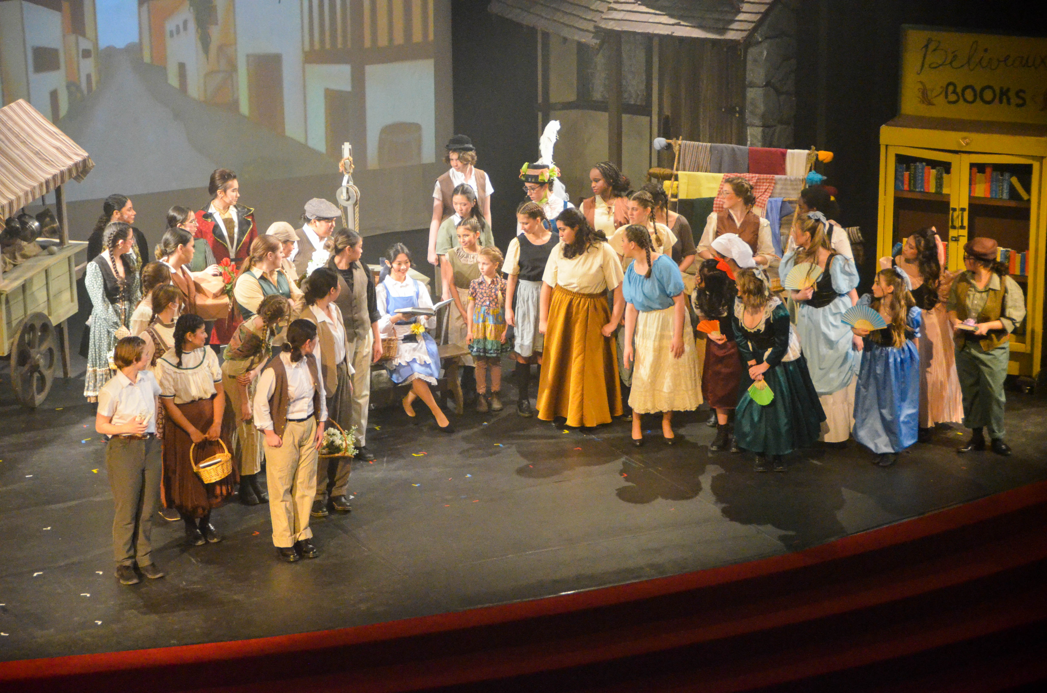 Students perform a scene from Disney's Beauty and the Beast, with the character Belle sitting with an open book surrounded by townspeople.