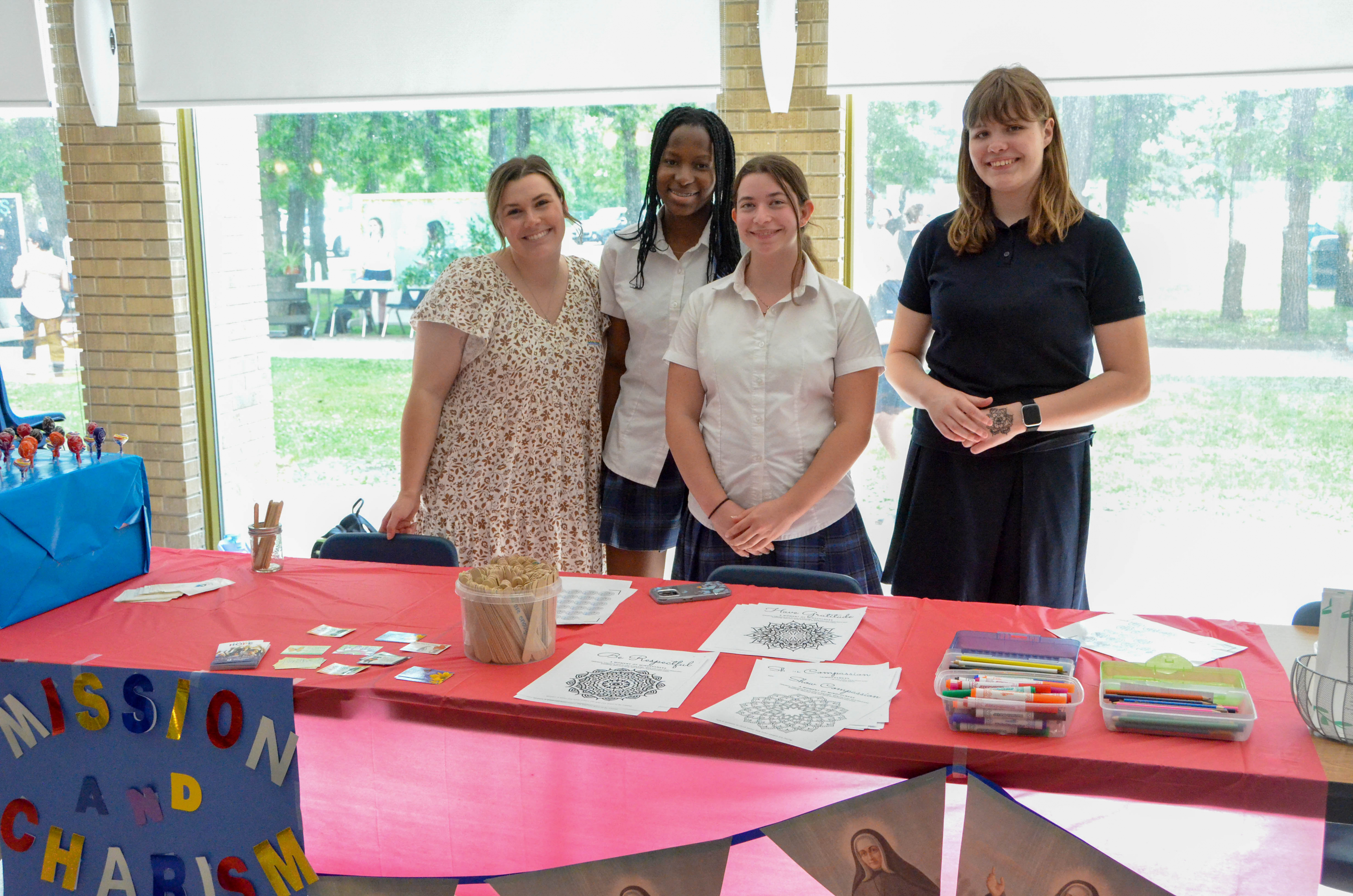 Four students stand behind a table with a red tablecloth with a 'Mission and Charism' sign on the front.