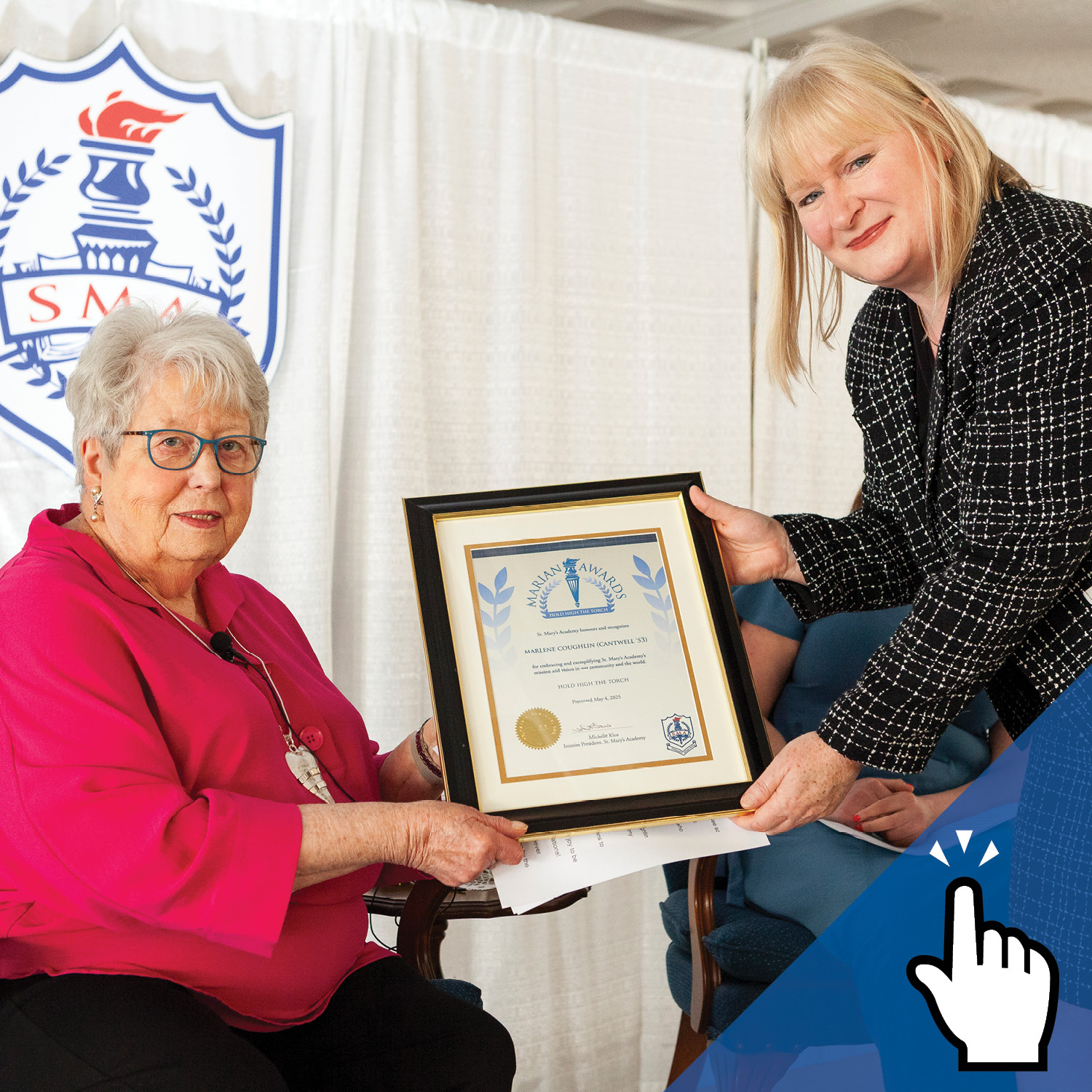 One woman sitting and receiving a framed award certificate from another woman standing.
