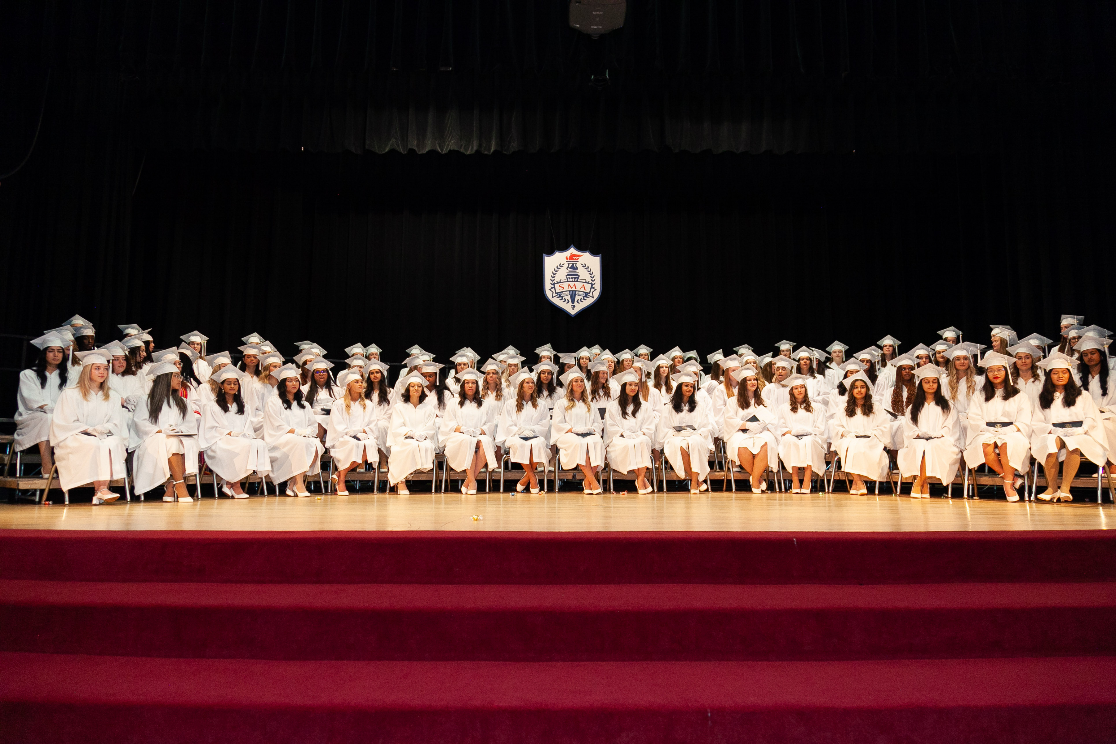 St. Mary's Academy graduates in caps and gowns sit on stage with a dark backdrop that features the Academy's crest.