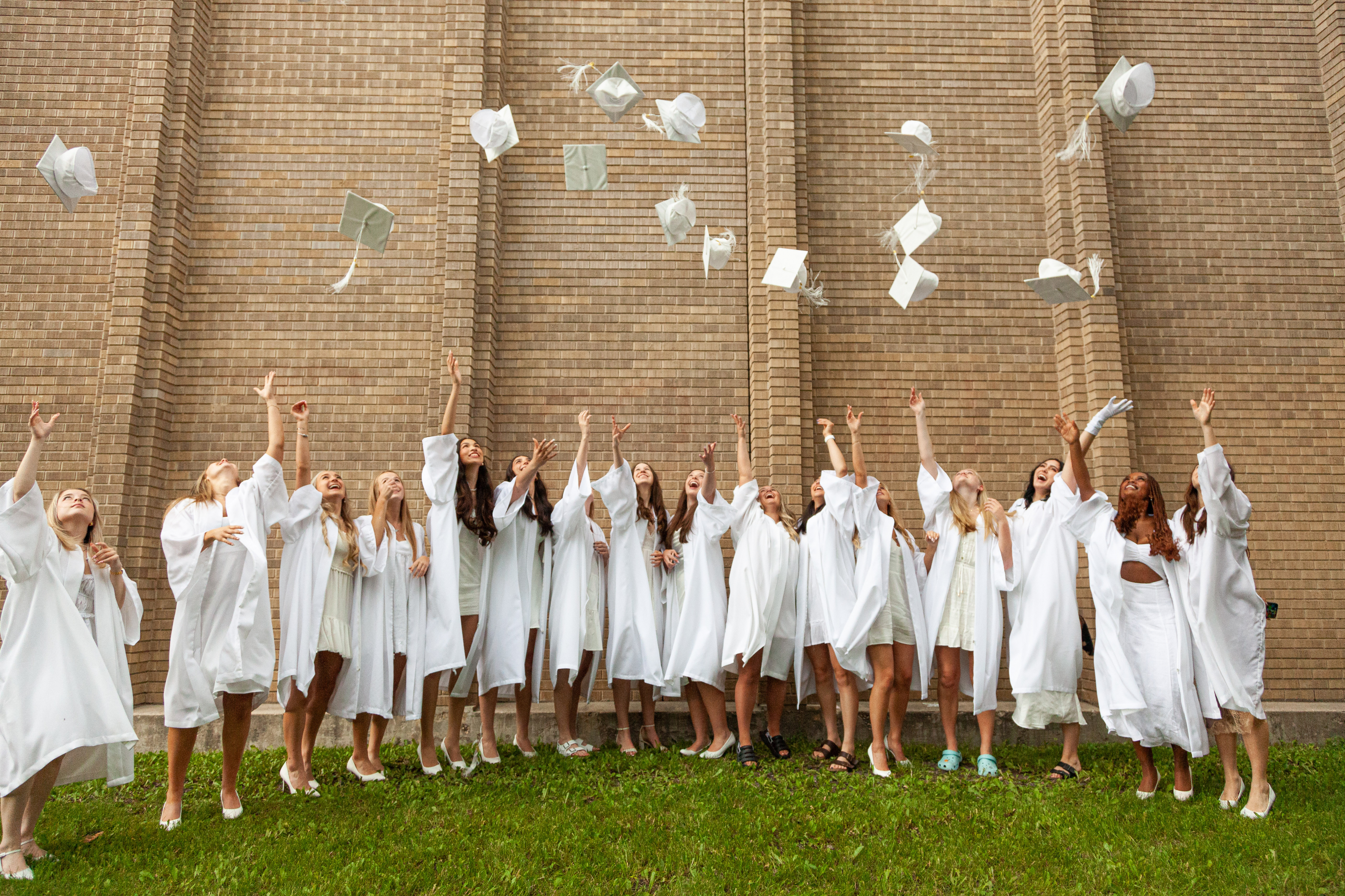 St. Mary's Academy graduates in gowns throw their caps in the air in front of a brick wall outside.