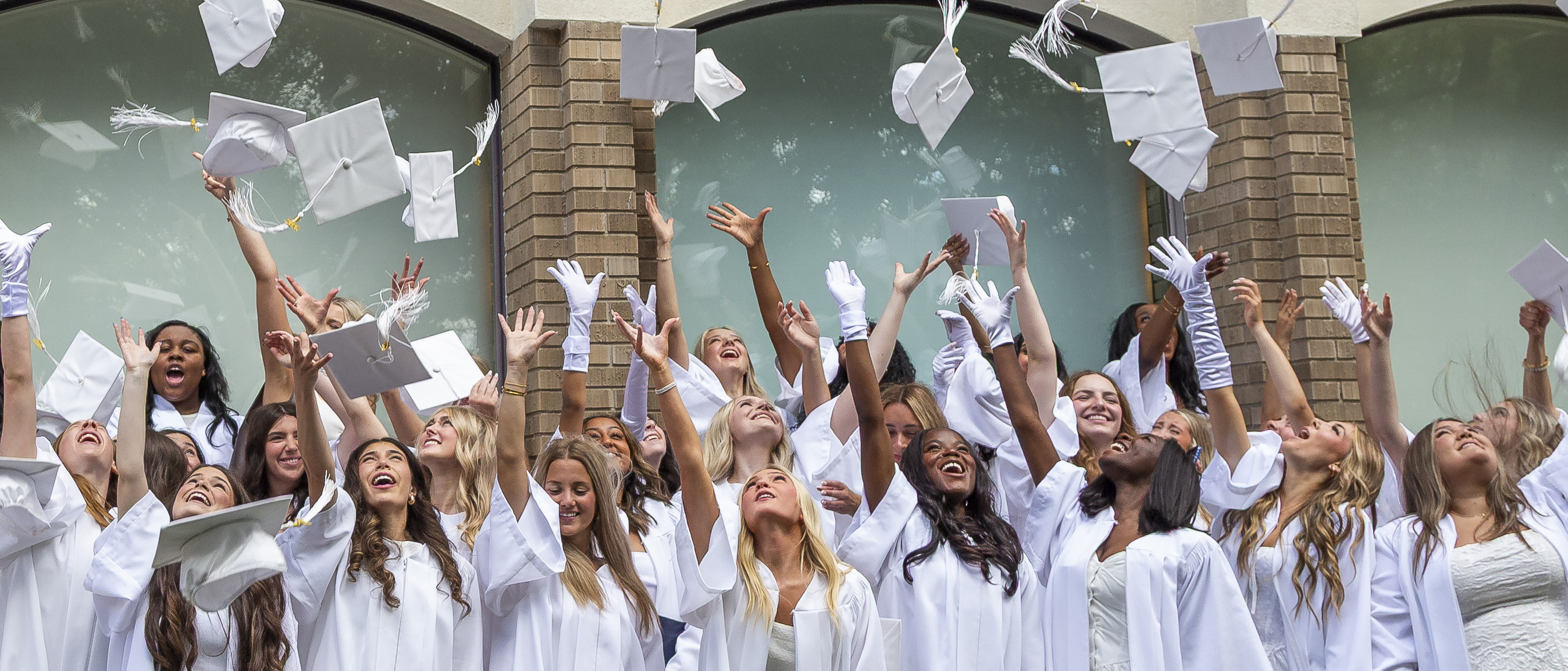 a group of girl students wearing white graduation gowns throwing their graduation caps into the air