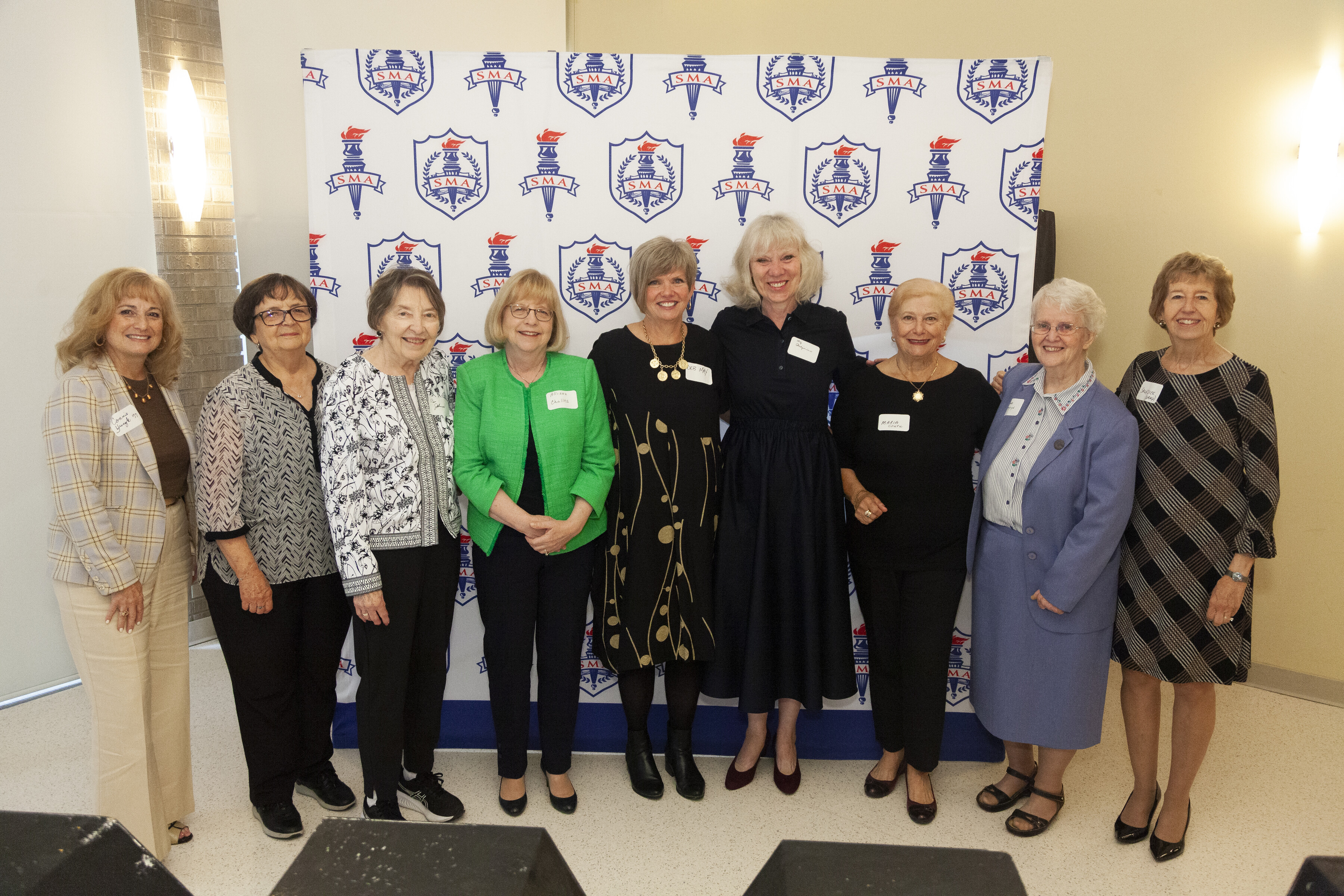 Nine smiling former SMA faculty and staff stand in front of a St. Mary's Academy backdrop.