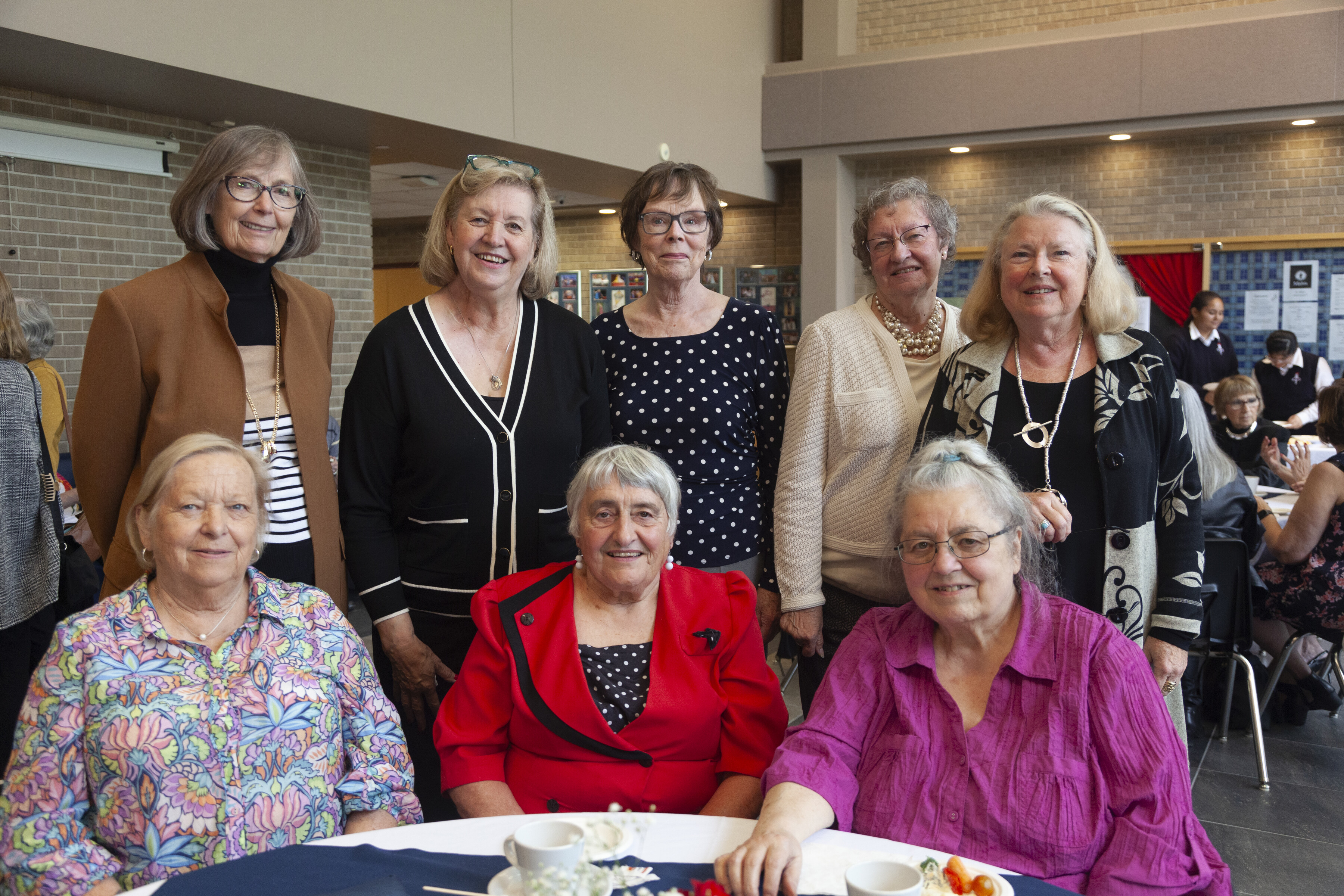 Seven smiling alumnae stand and sit at a table in the St. Mary's Academy atrium.