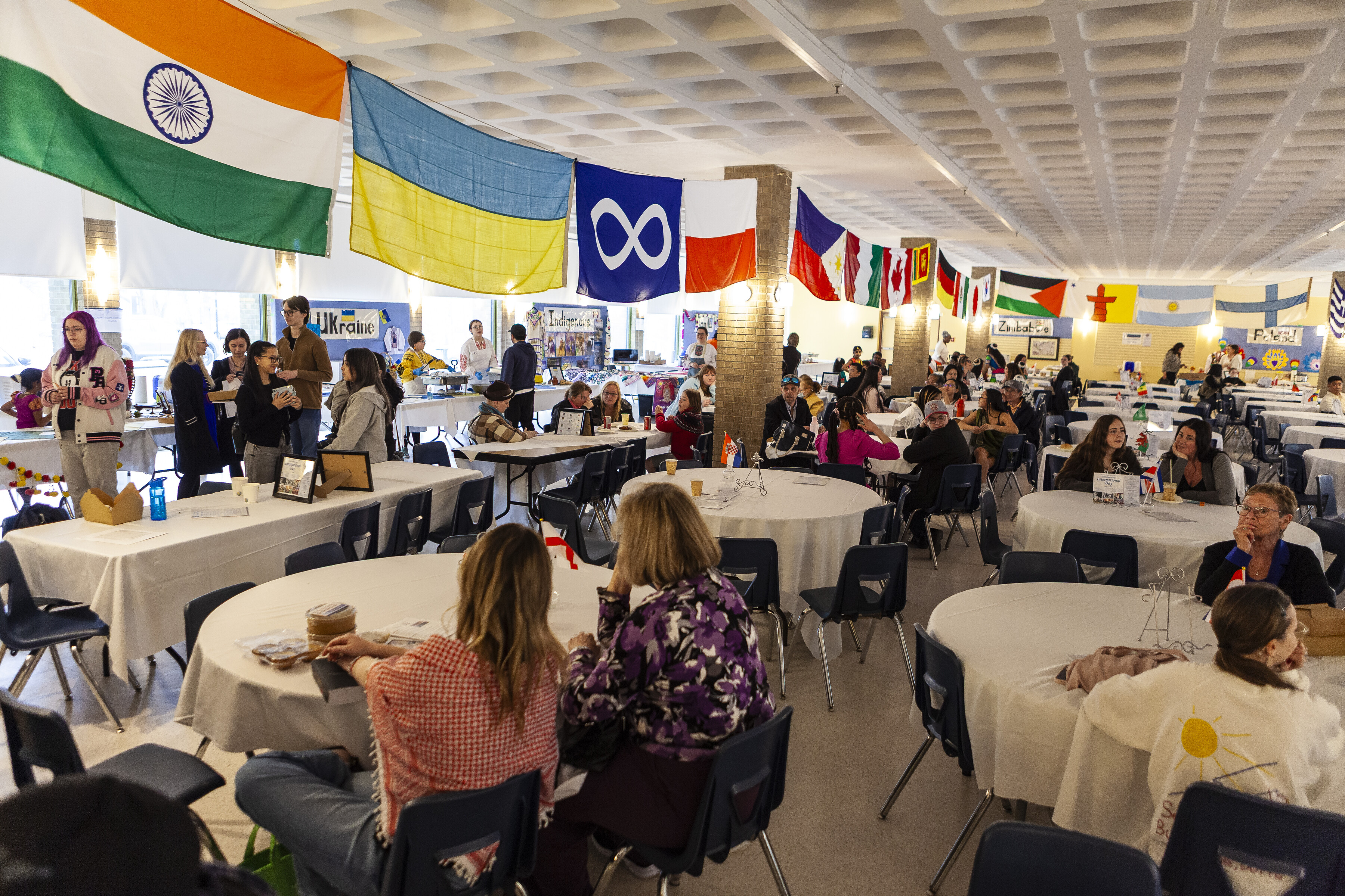 A full room of guests sit at tables during St. Mary's Academy International Day, with flags from around the world hanging from the ceiling.