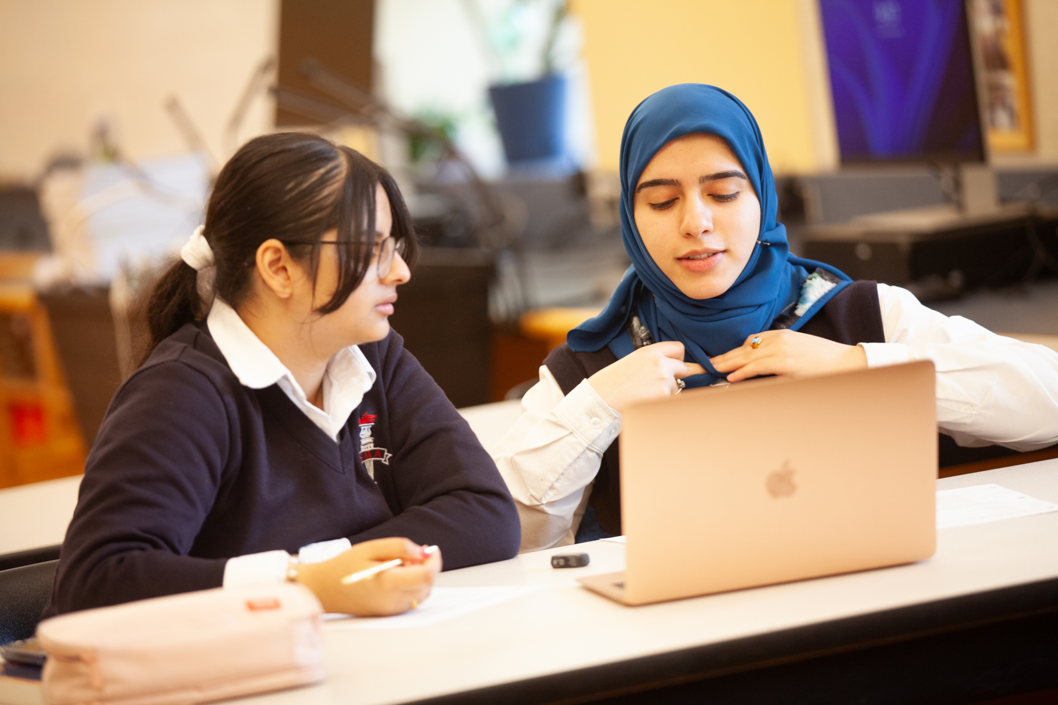 Two students sit at a desk in front of an open laptop.