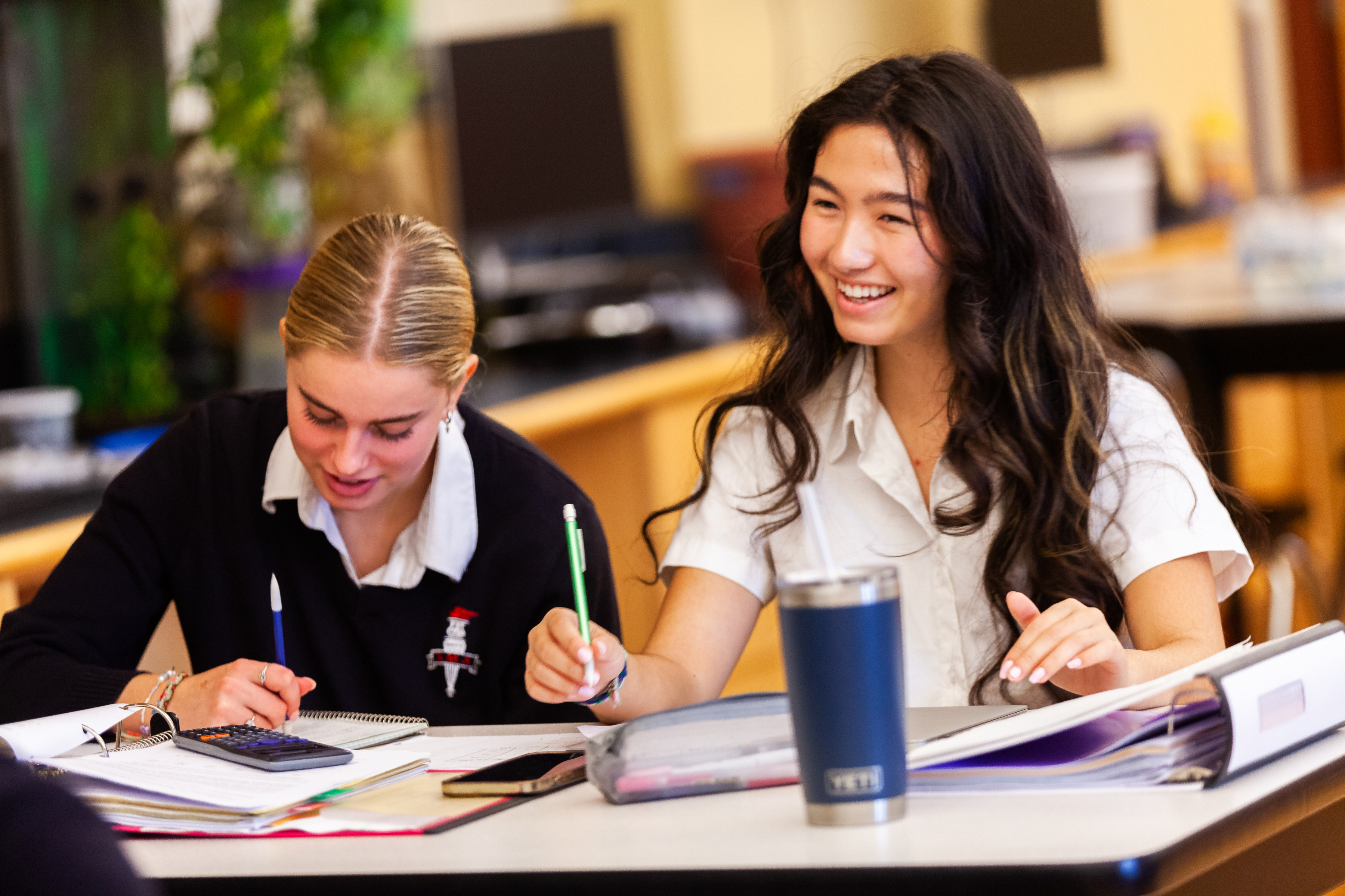 Two seated students take notes in a classroom, with a cup and calculator in front of them.