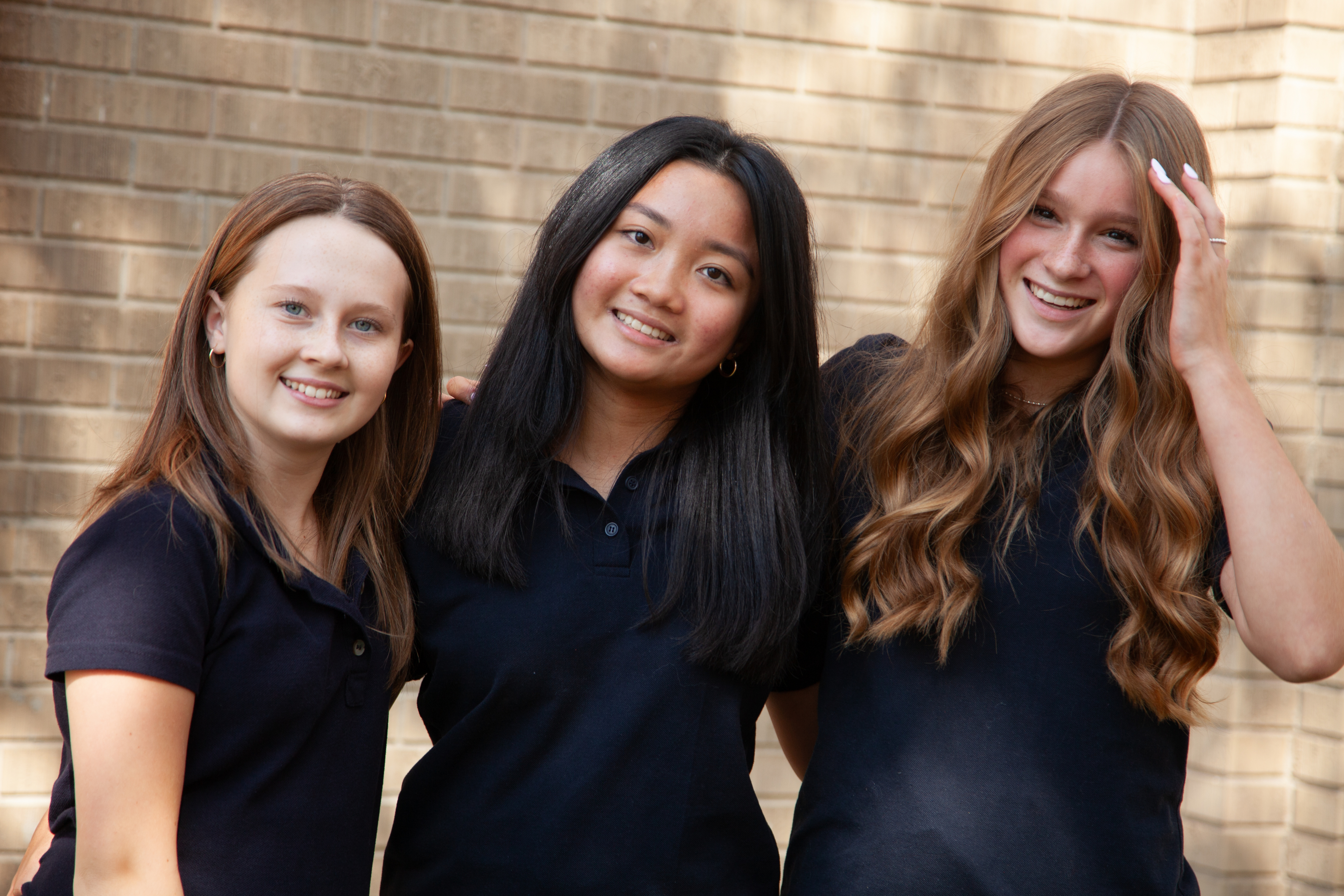 Three smiling students in black shirts stand in front of a brick wall.