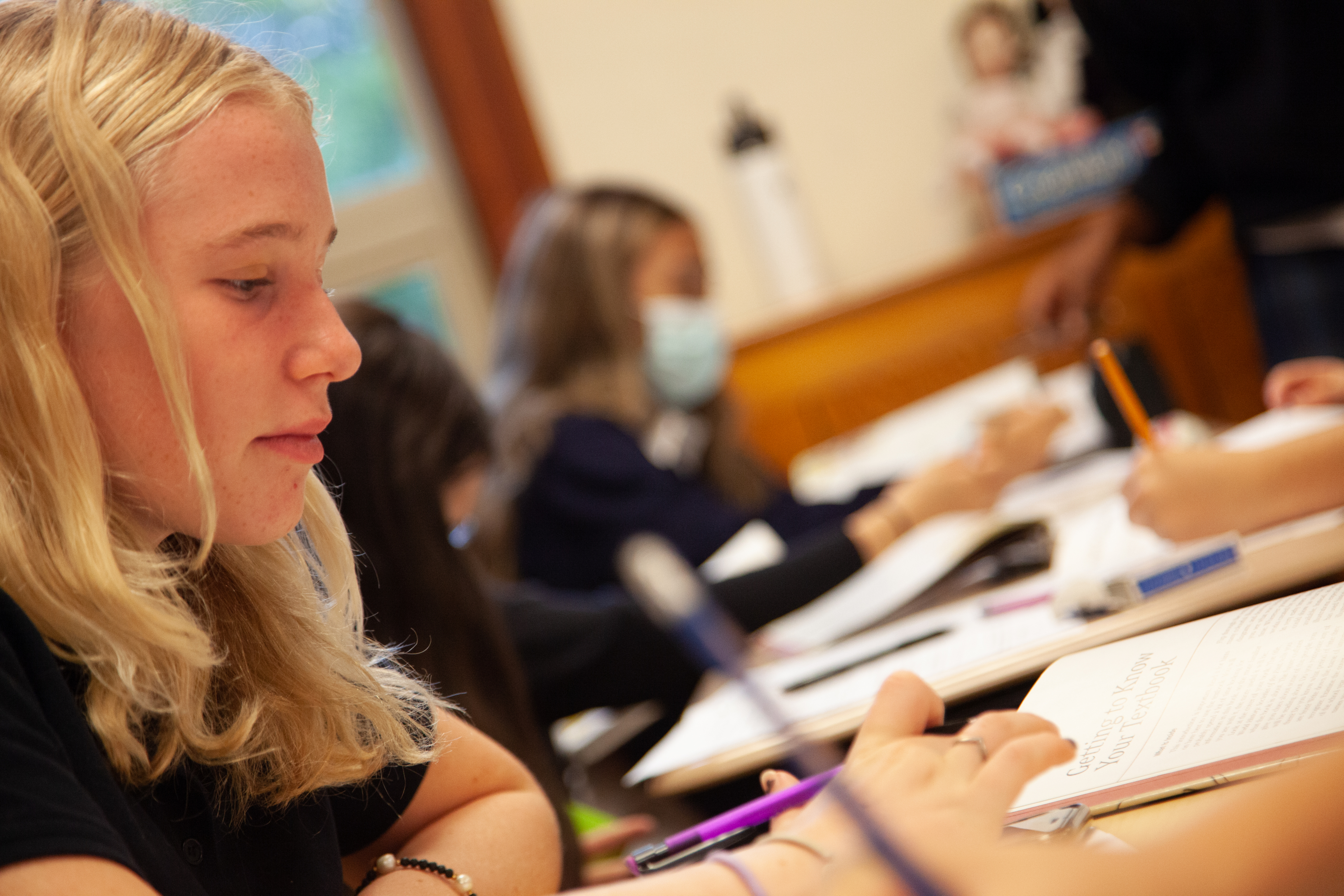A student writes with a pencil on a paper in a classroom, with other students doing the same in the background.