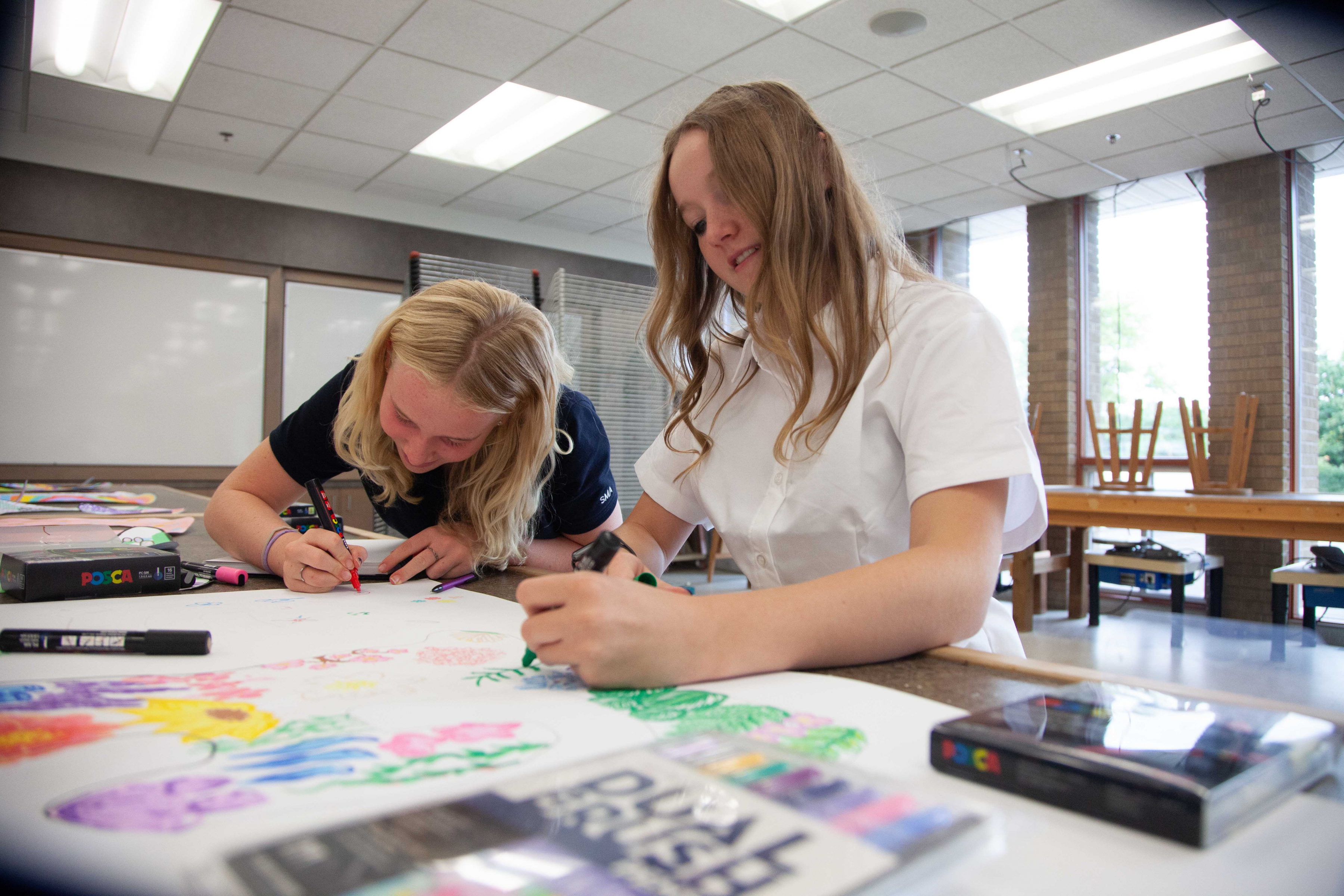 Two smiling St. Mary's Academy students colour a drawing with markers at a table.