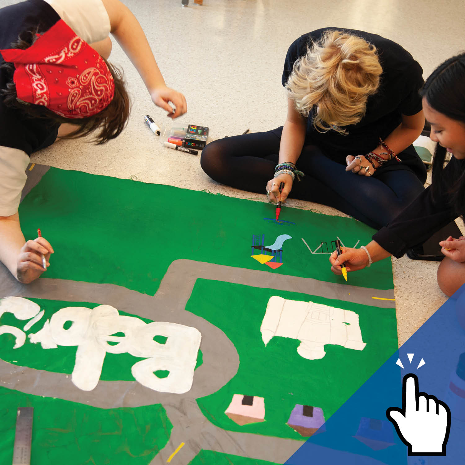 Three students sit on the ground and colour on a piece of art.