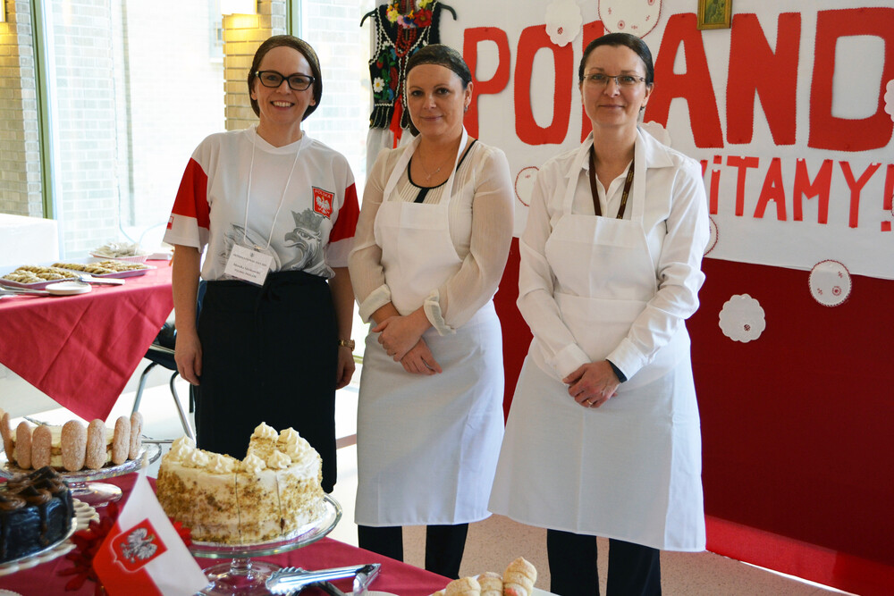 Three women smiling in front of a sign that says "Poland" with food placed on a table in the foreground
